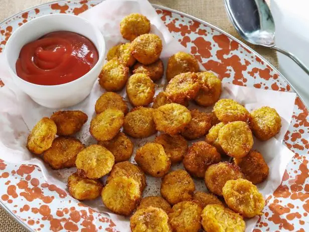 a luxurious spread of golden tater tots sit atop a plated, white napkin, delicately speckled w oil spots and fry crumbs, next to a small ramekin of perfectly swirled, fire engine red ketchup